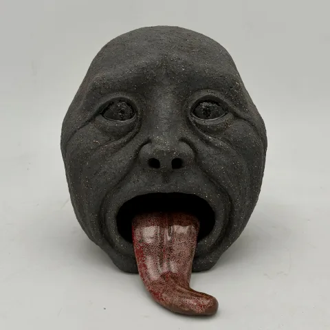 Dark speckled ceramic head with hollow eyes and a long glazed red tongue hanging from its open mouth.