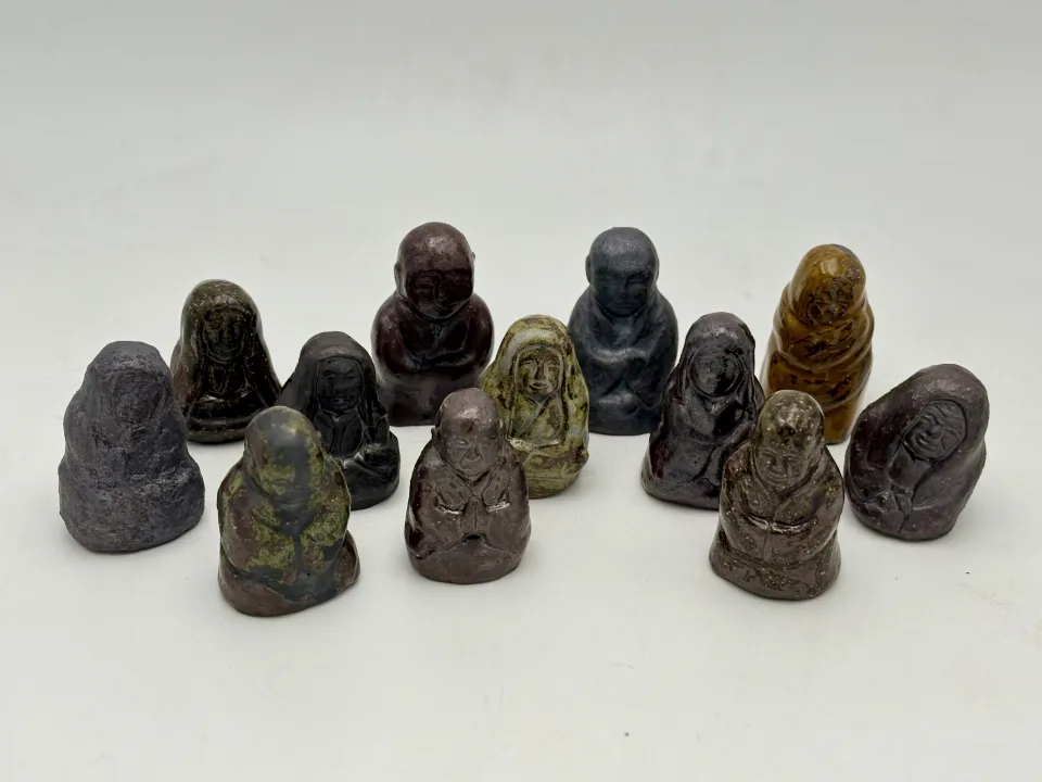 Twelve small glazed ceramic arhats in a loose group: robed and hooded figures in purple, grey, green and amber glazes.