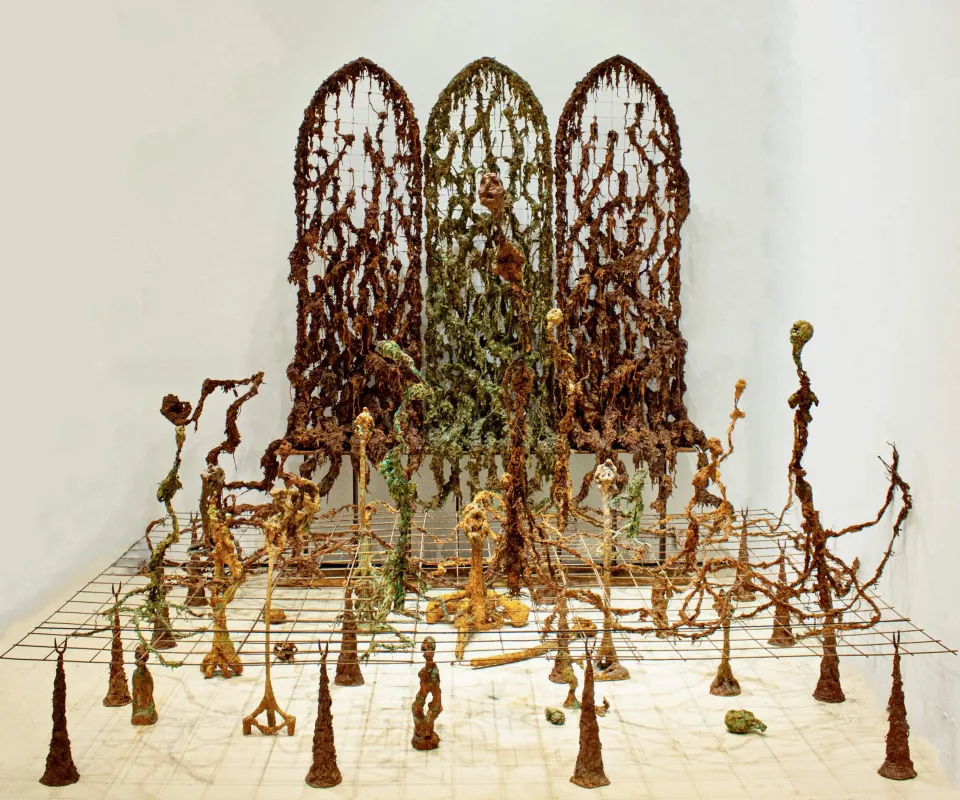 Installation of three arched lattice screens overgrown with root-like matter, standing over a grid field of thin sprouting figures.