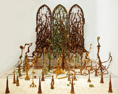 Installation of three arched lattice screens overgrown with rusted, root-like matter, standing over a grid floor from which thin, rust-coloured sprouting figures rise.