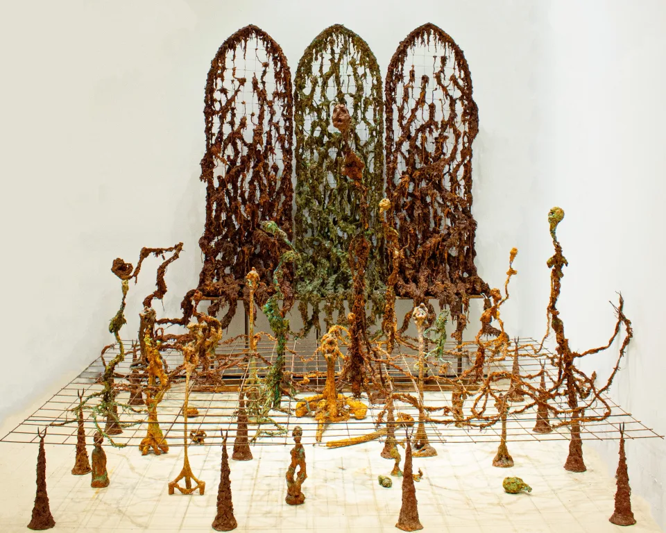 Installation of three arched lattice screens overgrown with rusted, root-like matter, standing over a grid floor from which thin, rust-coloured sprouting figures rise.