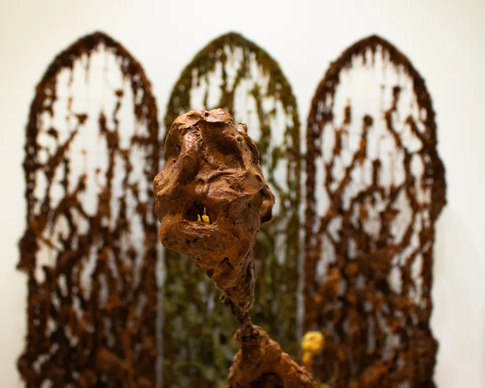 Detail: a rust-brown head-like form on a thin stem, the three screens blurred behind it.