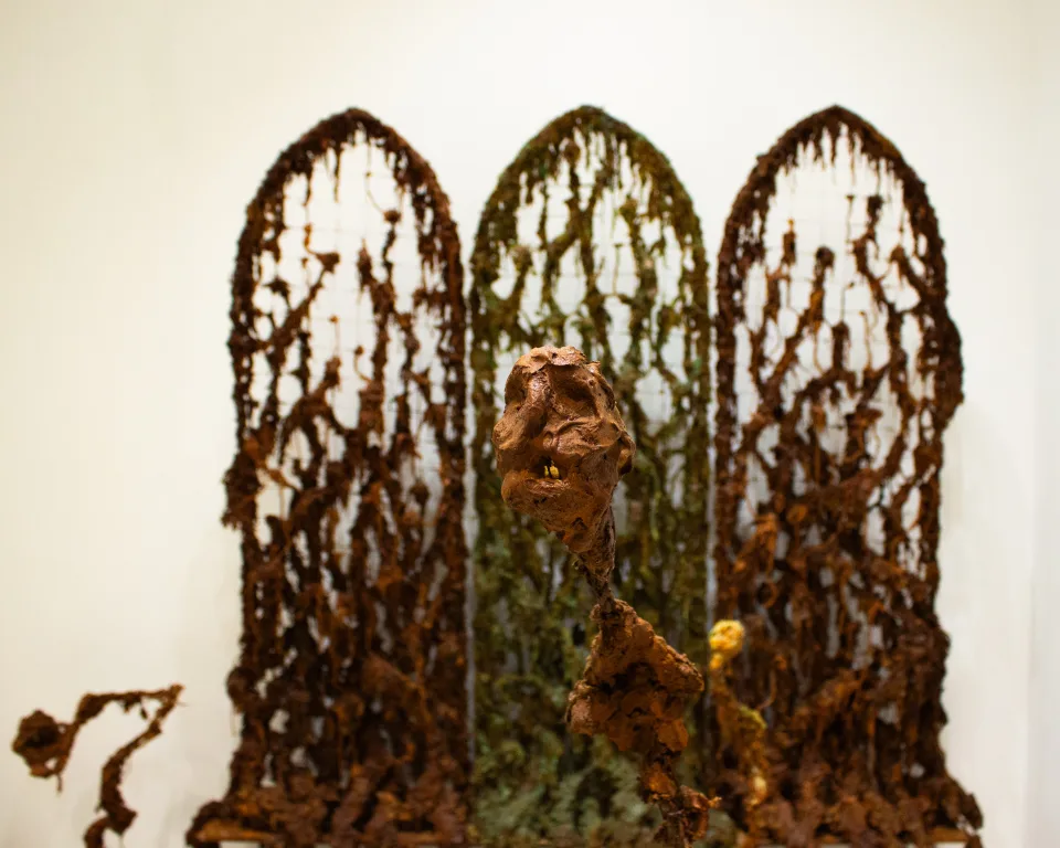 Detail: the head on its stem in front of the three screens, a small yellow figure at its foot.