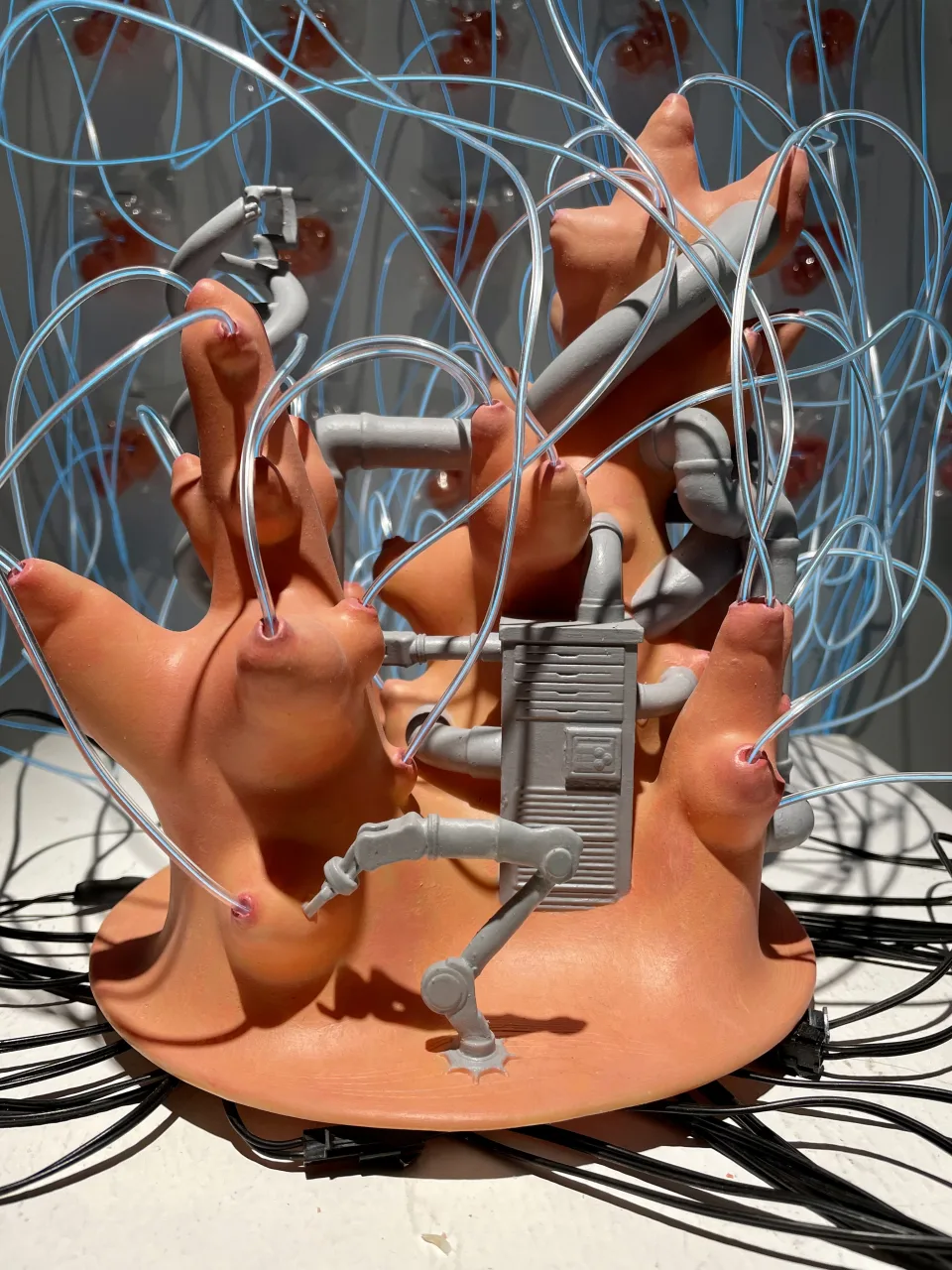 Detail: the central sculpture — fleshy orange forms tipped with pink nipples, grey pipes, a robotic arm and a ribbed box, tangled in blue wire.