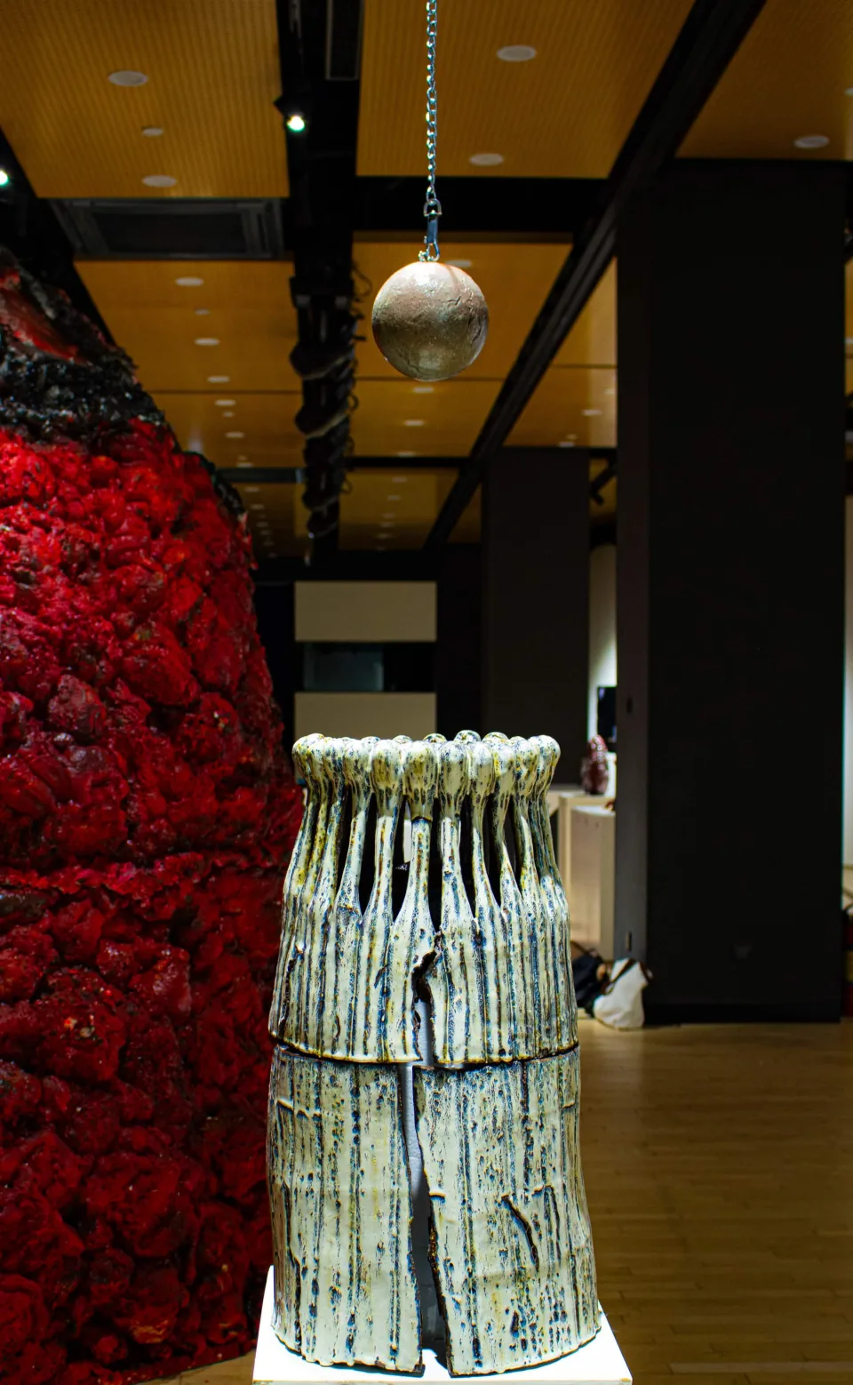 Tall glazed ceramic vessel with vertical slits standing on a plinth in an exhibition hall, a red mound and a hanging sphere behind it.