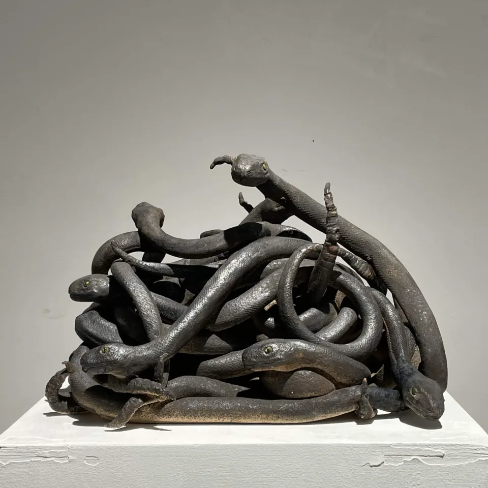 Ceramic sculpture of dark snakes tangled into a single mound, several heads emerging outward, on a white plinth.
