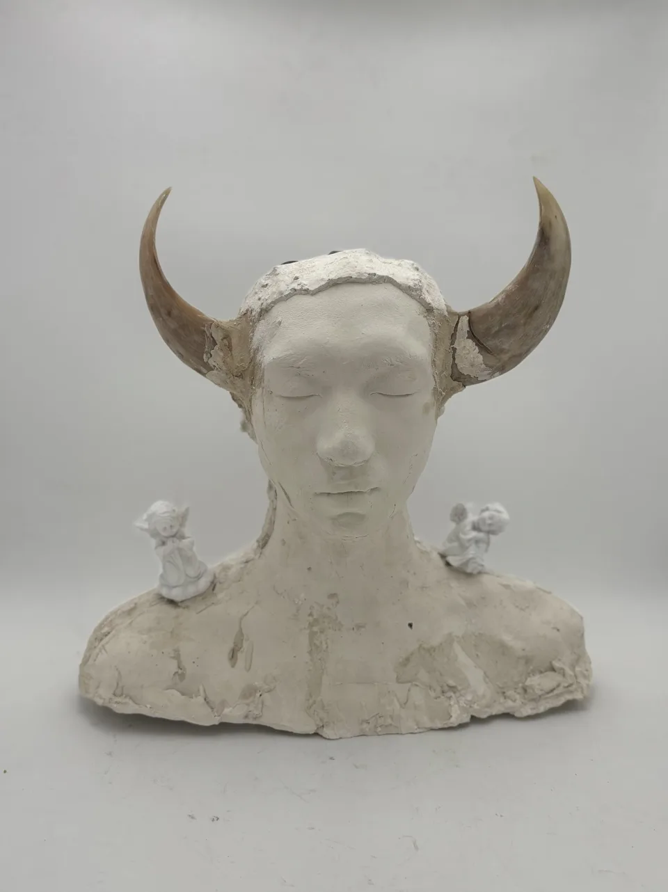 White plaster bust with closed eyes and two curved animal horns, a small white angel figurine on each shoulder.