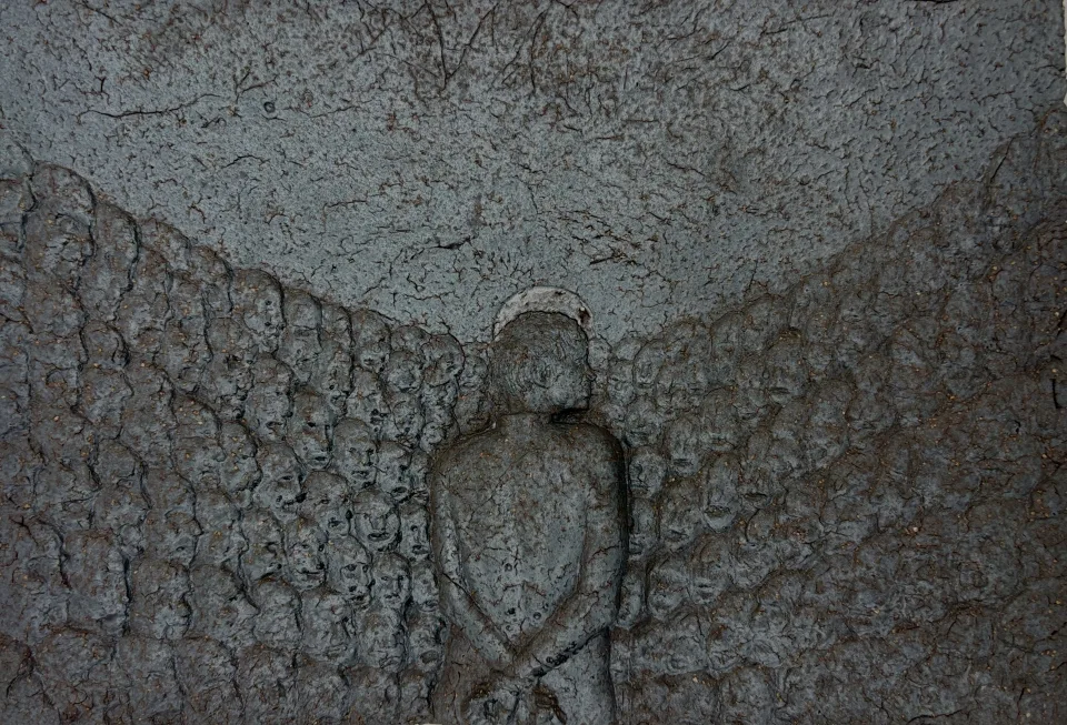 Glazed ceramic relief: a lone figure seen from behind, surrounded by a field of small anonymous faces.