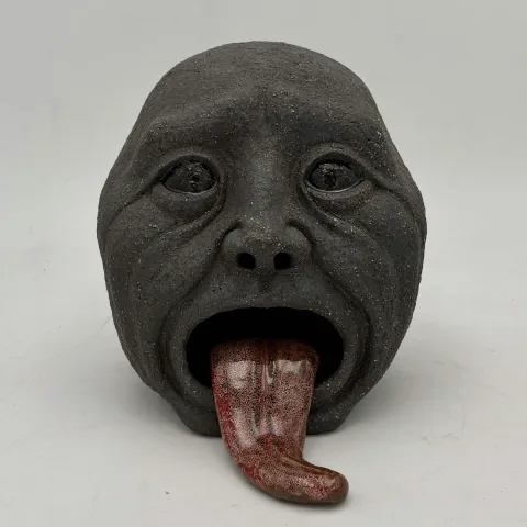 Dark speckled ceramic head with hollow eyes and a long glazed red tongue extending from its open mouth.